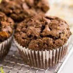 A close-up of chocolate zucchini muffins with chocolate chips, wrapped in a white paper liner, resting on a cooling rack.