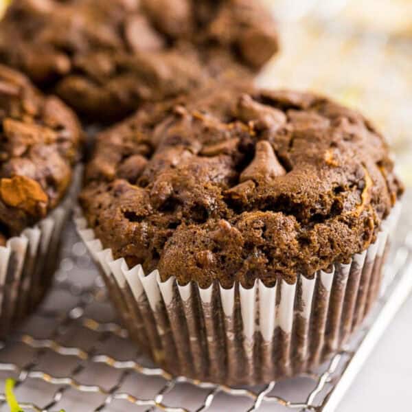 A close-up of chocolate zucchini muffins with chocolate chips, wrapped in a white paper liner, resting on a cooling rack.