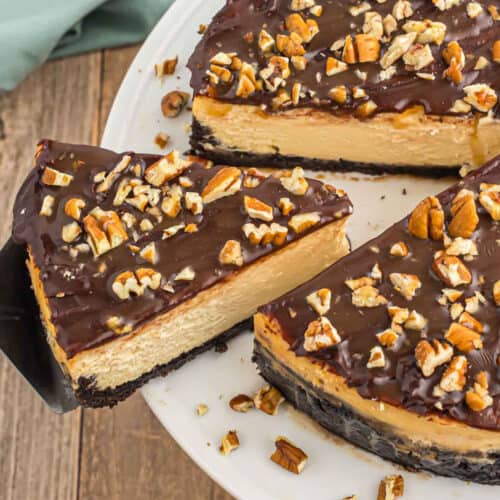 A sliced Turtle Cheesecake with a chocolate crust, topped with a glossy chocolate layer and chopped pecans, is shown on a white plate with one decadent piece being lifted.