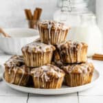A plate of Cinnamon Roll Muffins stacked in a pyramid, topped with white icing drizzle. In the background are a glass of milk, a jar, and cinnamon sticks. The scene is bright and inviting.
