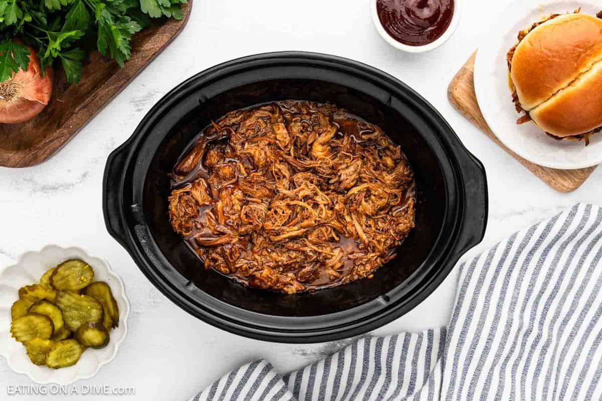 Pulled Pork in a crock pot with pickles and BBQ sauce in a bowl with a sandwich on the side. 