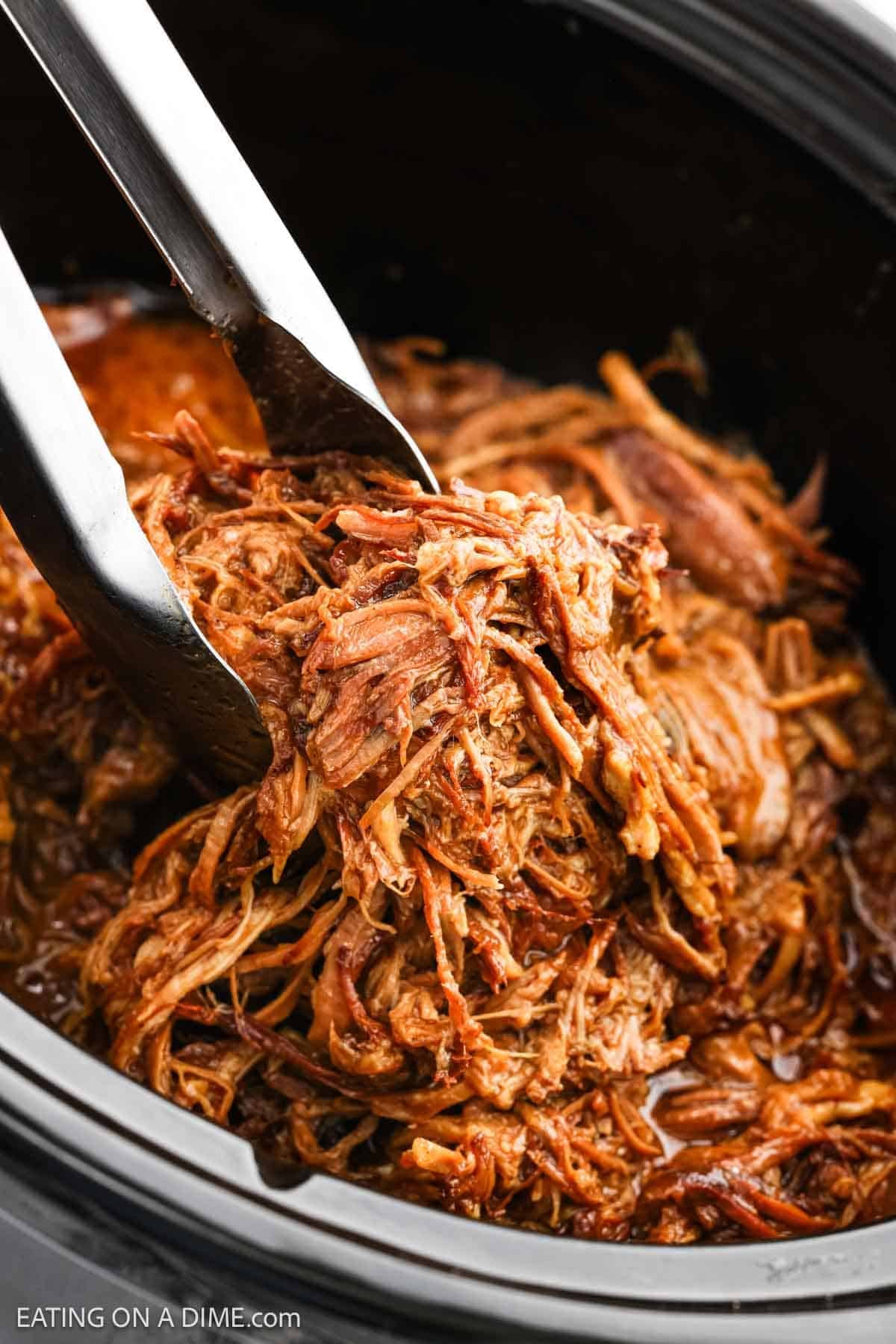 Shredded pork in a crock pot with a serving gathered with tongs.