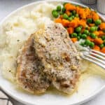 Crockpot Ranch Pork Chops with creamy gravy rest on mashed potatoes, accompanied by carrots and green peas on a white plate, with a fork on the side.