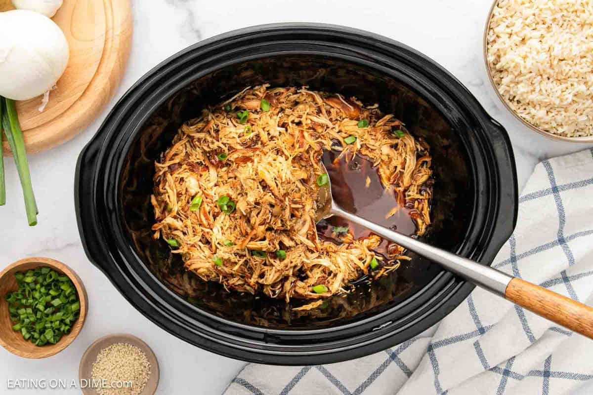 A crock pot teriyaki chicken recipe simmers in a slow cooker, filled with shredded chicken in a dark sauce and topped with green onions. A ladle rests inside, with bowls of rice, sesame seeds, and extra green onions nearby on a cloth napkin.