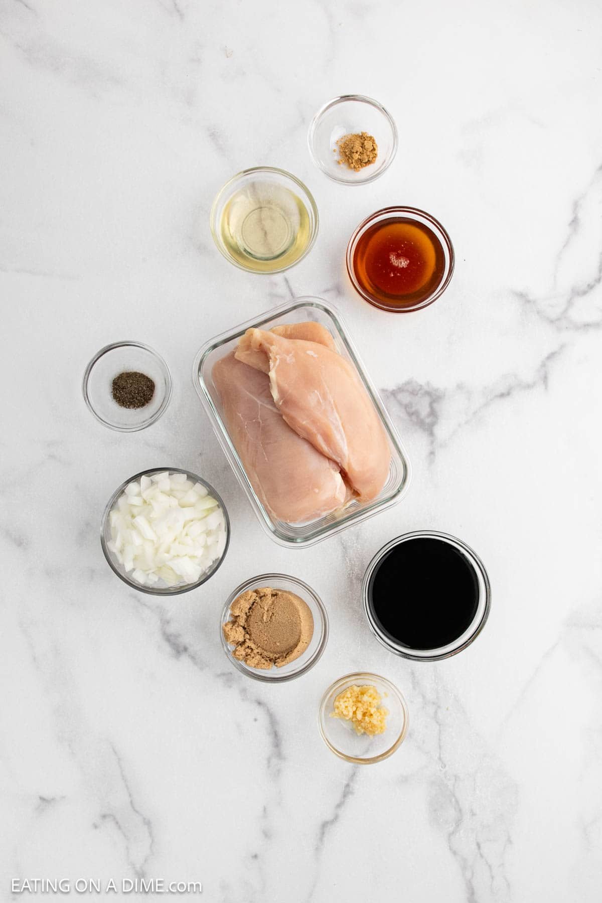 Overhead view of raw chicken breasts in a glass container, surrounded by small bowls of ingredients for a crock pot teriyaki chicken recipe—chopped onions, brown sugar, minced garlic, black pepper, soy sauce, honey, ground ginger, and rice vinegar.