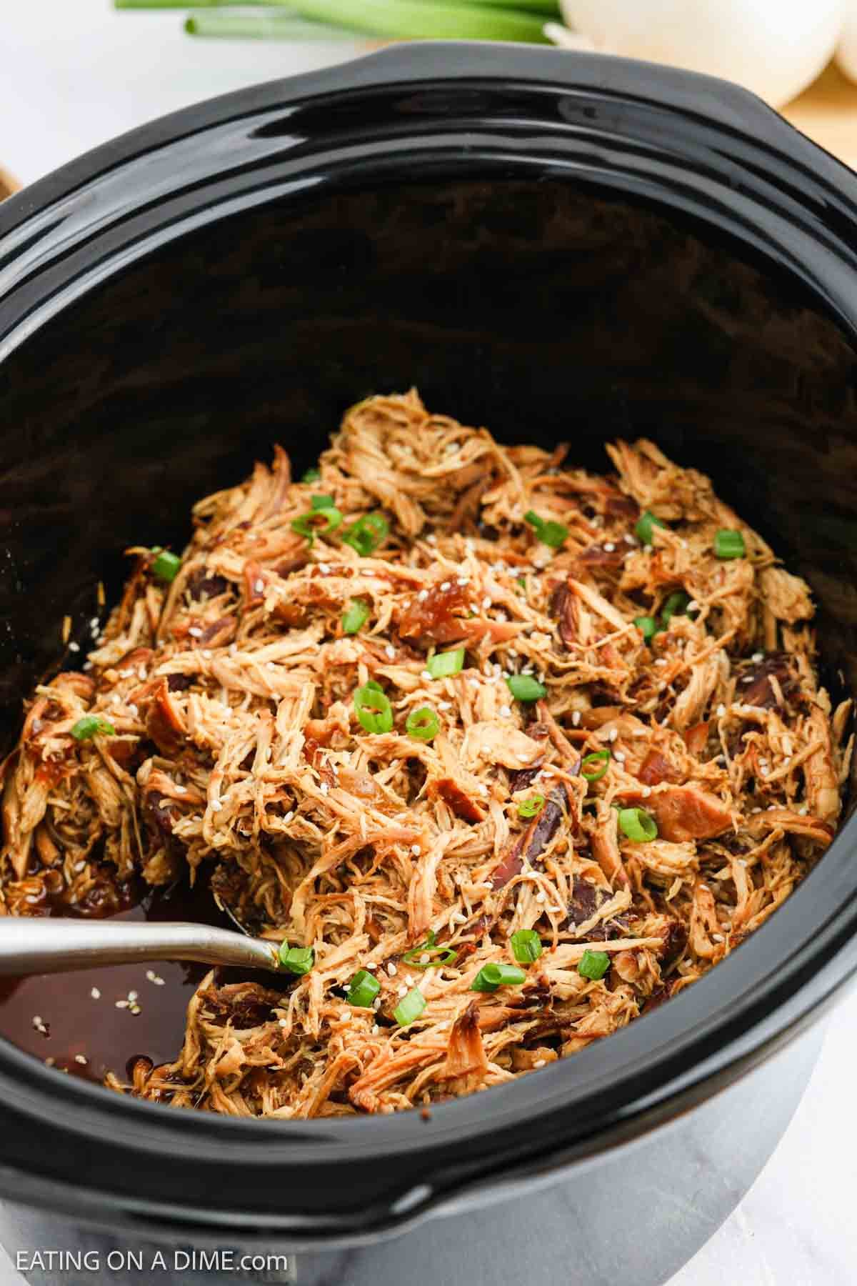 Shredded chicken with teriyaki sauce, garnished with chopped green onions and sesame seeds, served in a black crock pot with a metal serving spoon—an easy crock pot teriyaki chicken recipe perfect for any meal.
