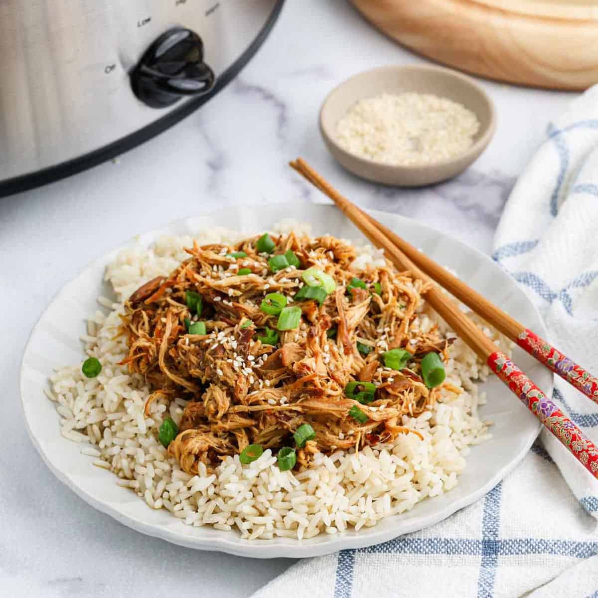 A plate of white rice topped with shredded crock pot teriyaki chicken, garnished with sesame seeds and chopped green onions. Chopsticks rest on the plate, alongside a small bowl of sesame seeds and a slow cooker.