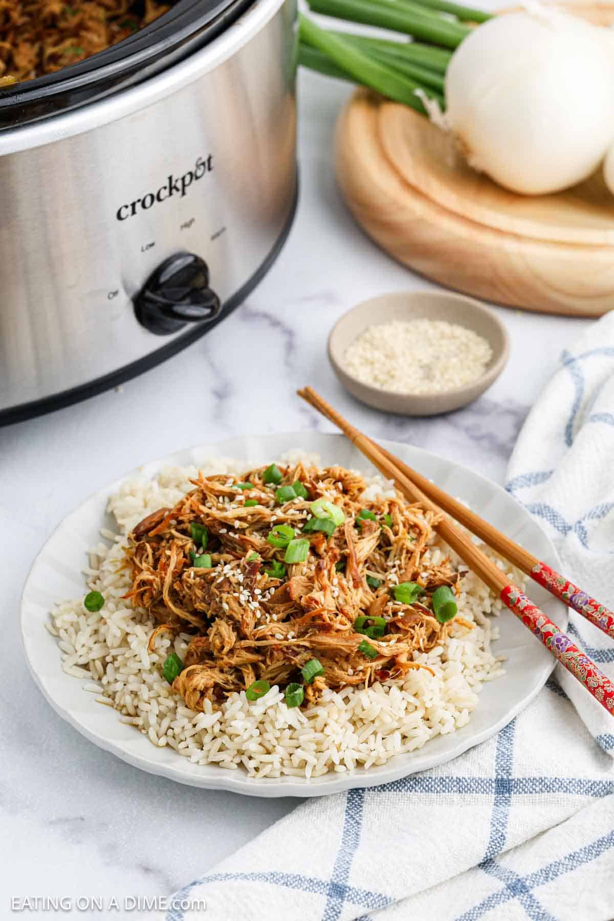 A plate of shredded crock pot teriyaki chicken recipe in sauce served over white rice, garnished with chopped green onions and sesame seeds. Chopsticks rest on the side, with a Crockpot, green onions, and a whole onion in the background.