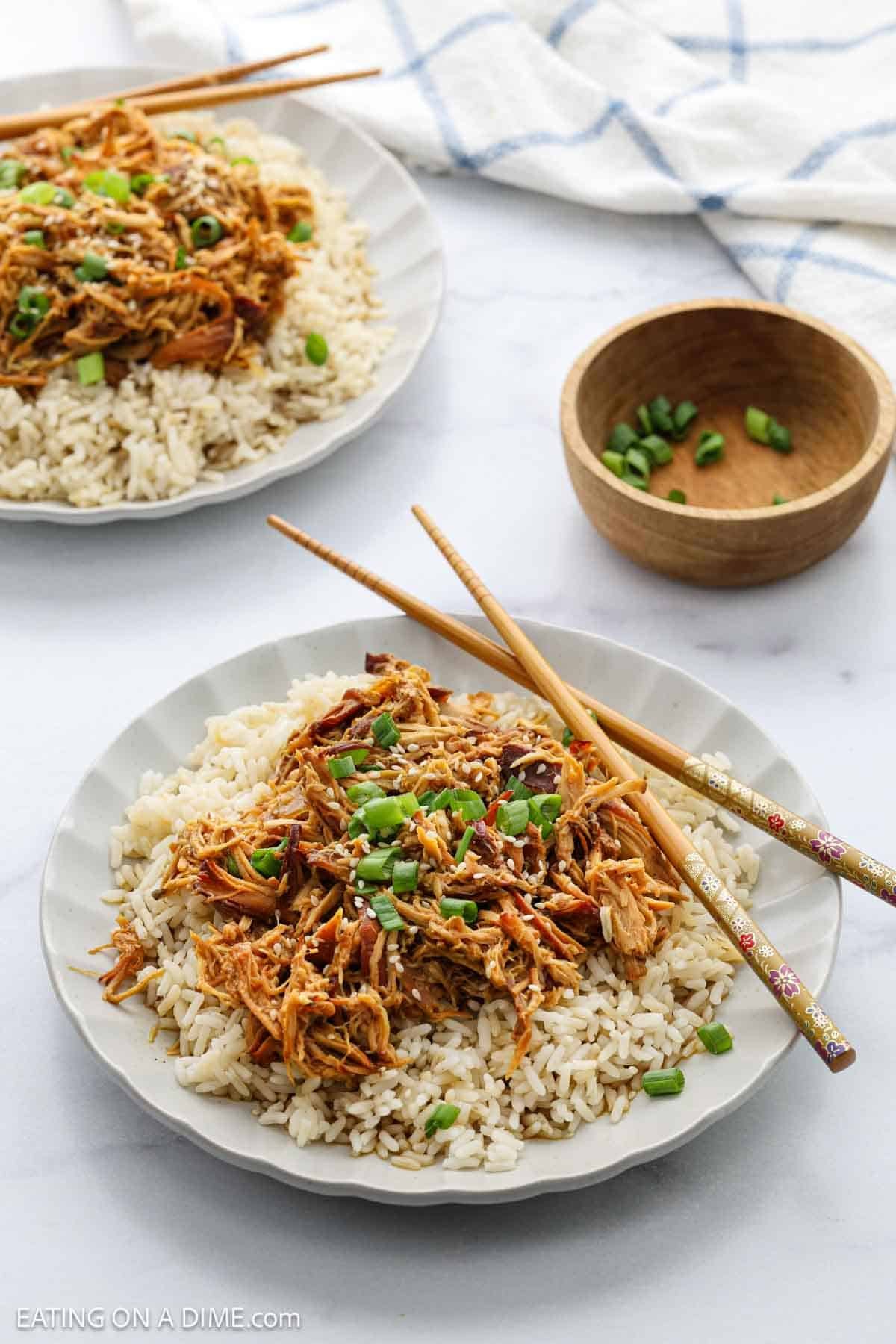 Two plates of crock pot teriyaki chicken recipe are served over white rice, garnished with chopped green onions. Wooden chopsticks rest on the plates, with a small bowl of extra green onions nearby on a white surface.