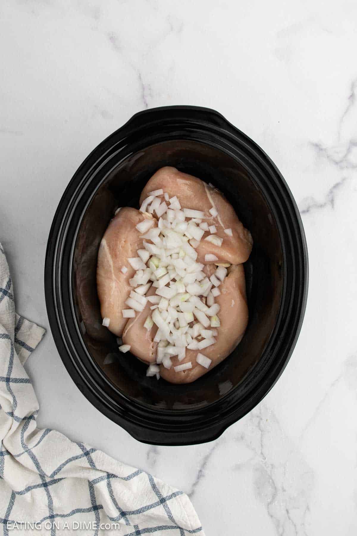A black slow cooker with raw chicken breasts topped with chopped onions, ready for a crock pot teriyaki chicken recipe, sits on a white marble surface next to a white and blue striped cloth.
