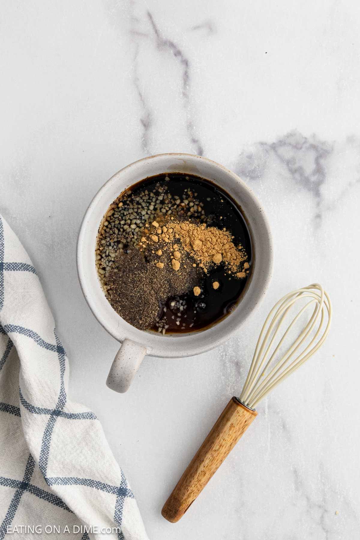A white bowl filled with dark liquid and spices, likely part of a crock pot teriyaki chicken recipe, sits next to a small whisk with a wooden handle on a light marble surface beside a blue and white checkered cloth.