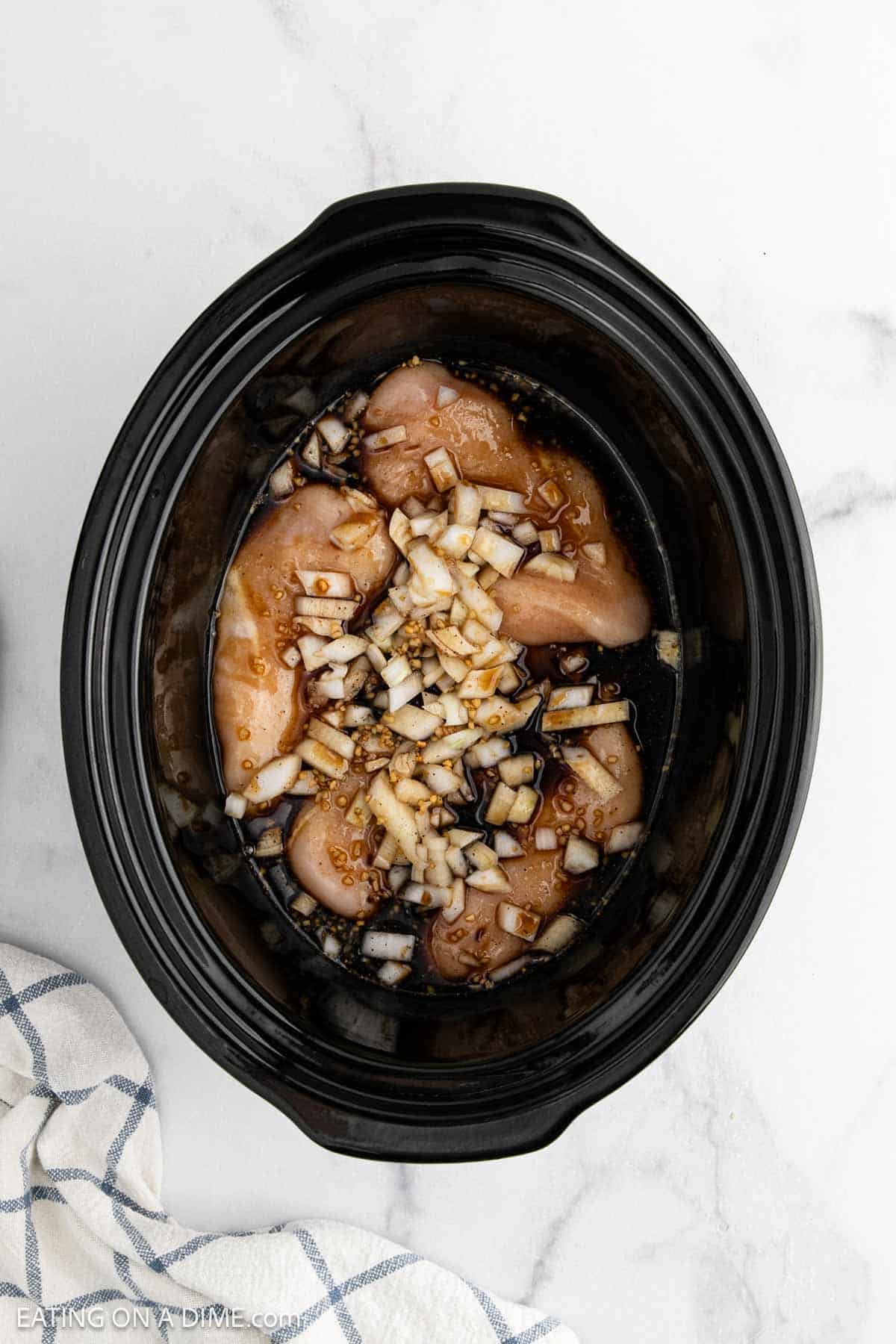 Raw chicken breasts, chopped onions, and sauce ingredients are ready in a black slow cooker for an easy crock pot teriyaki chicken recipe. The slow cooker sits on a white surface beside a striped kitchen towel.