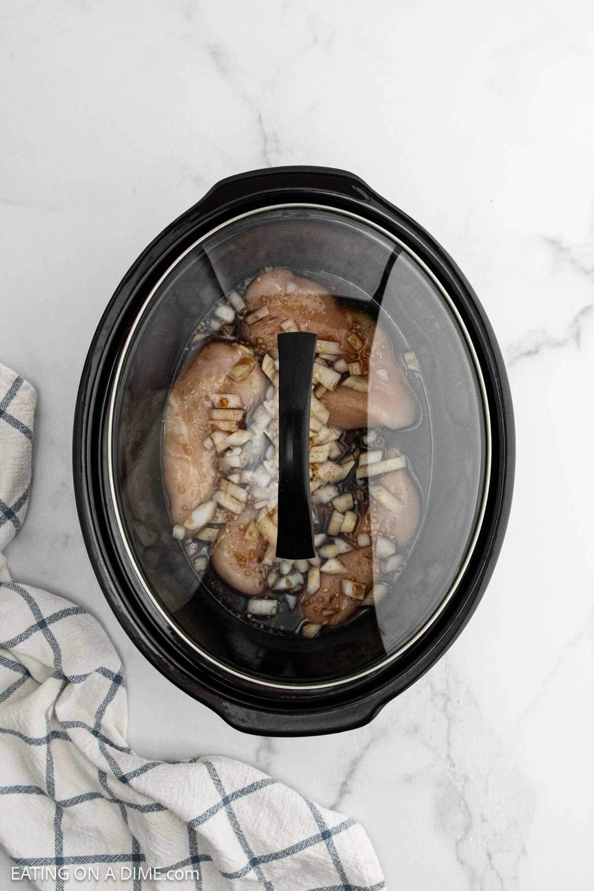 Overhead view of a crock pot teriyaki chicken recipe, with raw chicken breasts, chopped onions, and sauce inside the slow cooker. The lid is on, and a white kitchen towel with blue stripes rests on a marble countertop nearby.