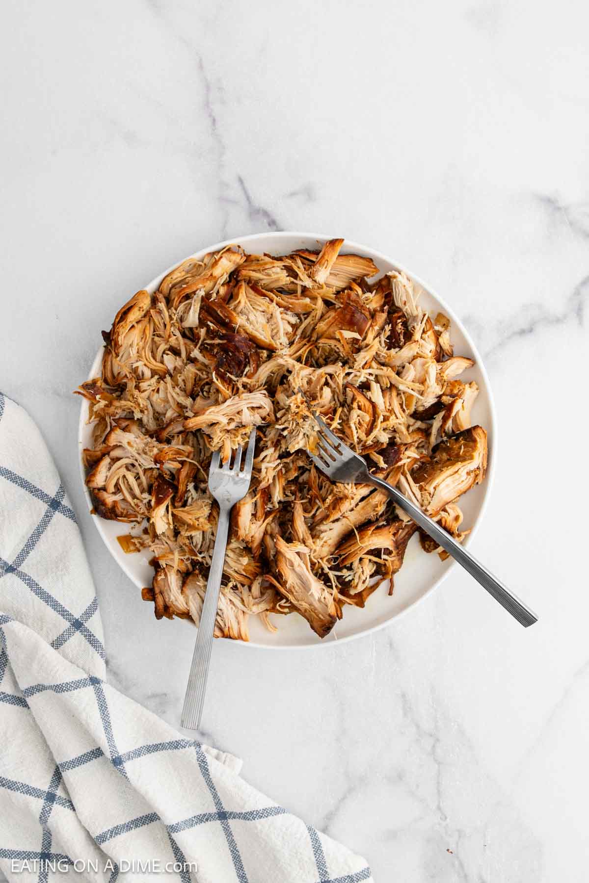 A white plate filled with shredded cooked chicken, prepared from a crock pot teriyaki chicken recipe, with two forks pulling apart the meat. The plate sits on a marble surface next to a blue and white checkered kitchen towel.