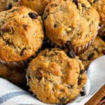 A close-up of several freshly baked peanut butter banana muffins with chocolate chips, stacked together in a basket lined with a white and blue striped cloth.