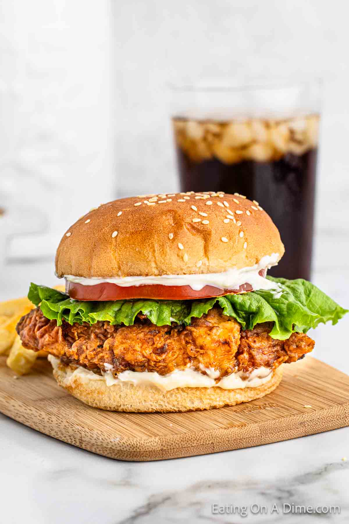 Crispy Chicken Sandwich topped with mayo, slice tomato, and lettuce with a sesame bun with a side of french fries and a glass of coca cola in the background