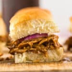 A close-up of a slider sandwich with tender Dr Pepper chicken and a slice of red onion inside a soft, golden-brown bun, resting on a wooden surface.