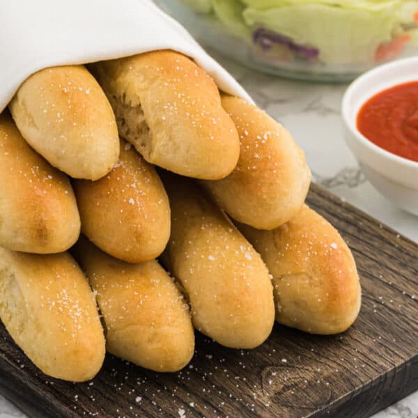 A stack of golden-brown breadsticks, inspired by the Olive Garden Breadsticks recipe, sprinkled with salt and partially covered by a white cloth, sits on a wooden board. In the background are bowls of red marinara sauce and fresh lettuce salad.
