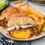 A Juicy Lucy Burger with lettuce, onions, and tomato on a sesame bun is cut in half on a plate, served with crinkle-cut fries and a pickle spear. Melted cheese oozes from the burger patty.