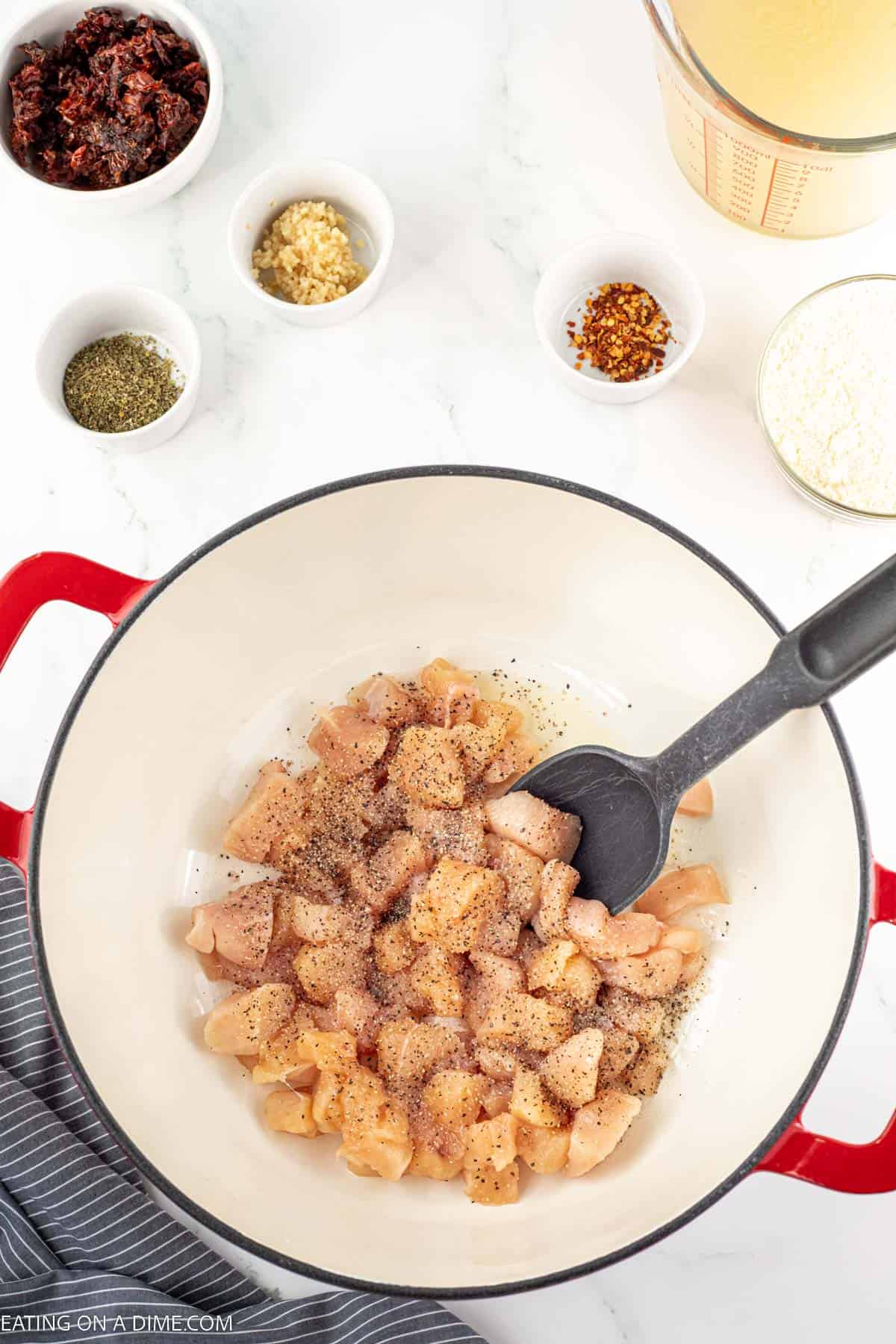 Diced Chicken in a large pot with seasoning on top with small bowls of seasoning and a measuring cup of chicken broth
