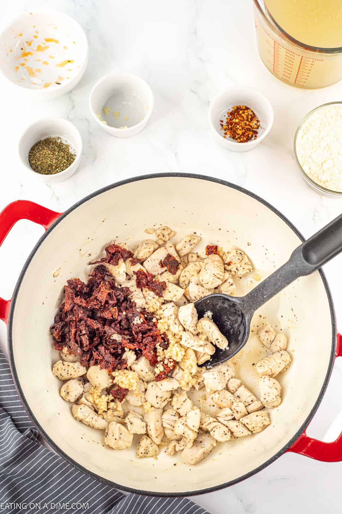Cooking diced chicken in a large pot topped with minced garlic and sun dried tomatoes with small bowls of seasoning on the side