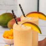 Two glasses of Mango Protein Smoothie with black polka dot straws, topped with shredded coconut and a mango slice, surrounded by fresh mango pieces and whole mangoes in the background.