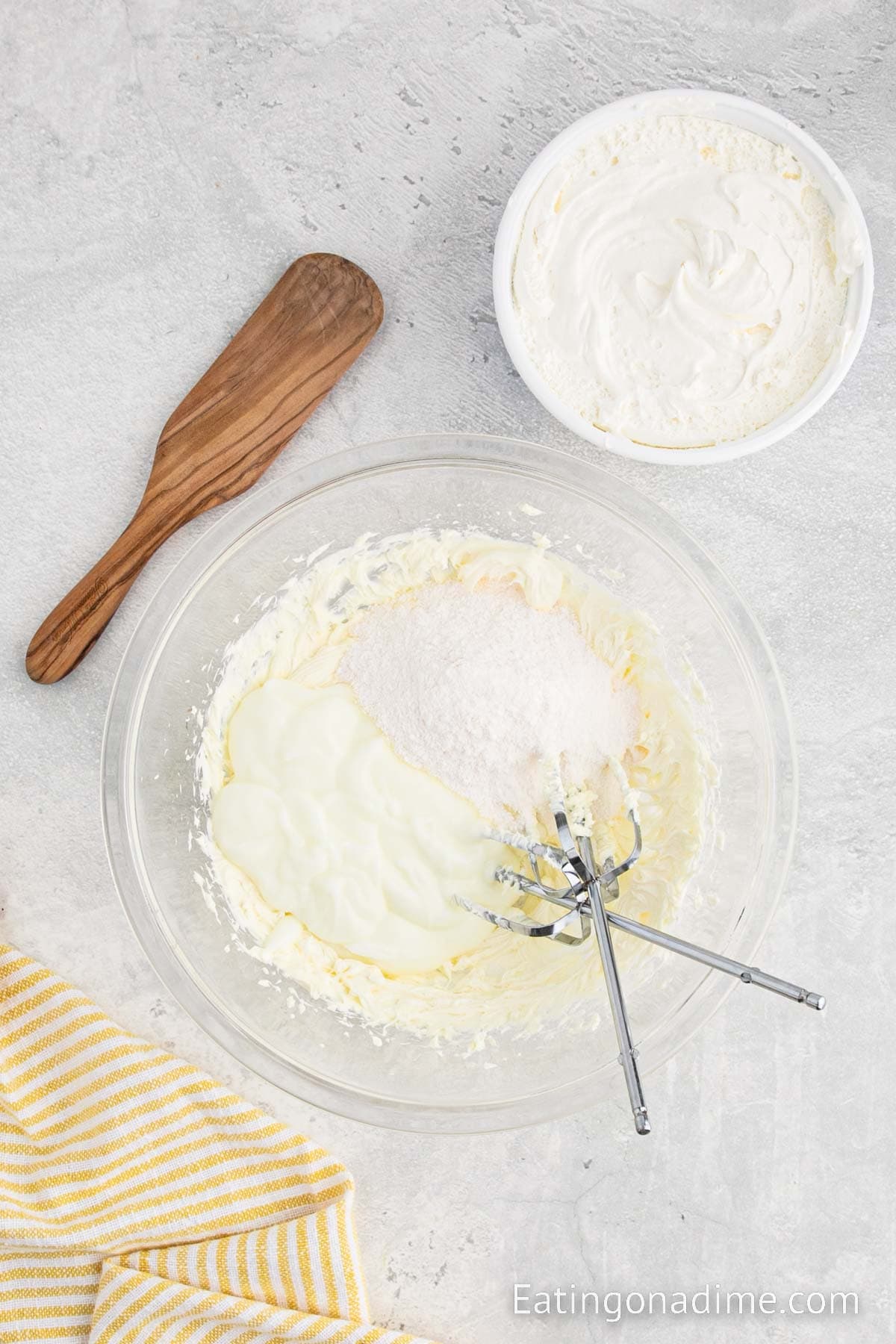 Mixing in the yogurt, pudding mix into the cream cheese mixture in a bowl with a container of cool whip on the side