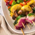 Colorful vegetable kabobs with mushrooms, zucchini, yellow squash, red onion, and bell peppers are arranged on a white platter. The seasoned vegetables are grilled to perfection and rest on a beige cloth.