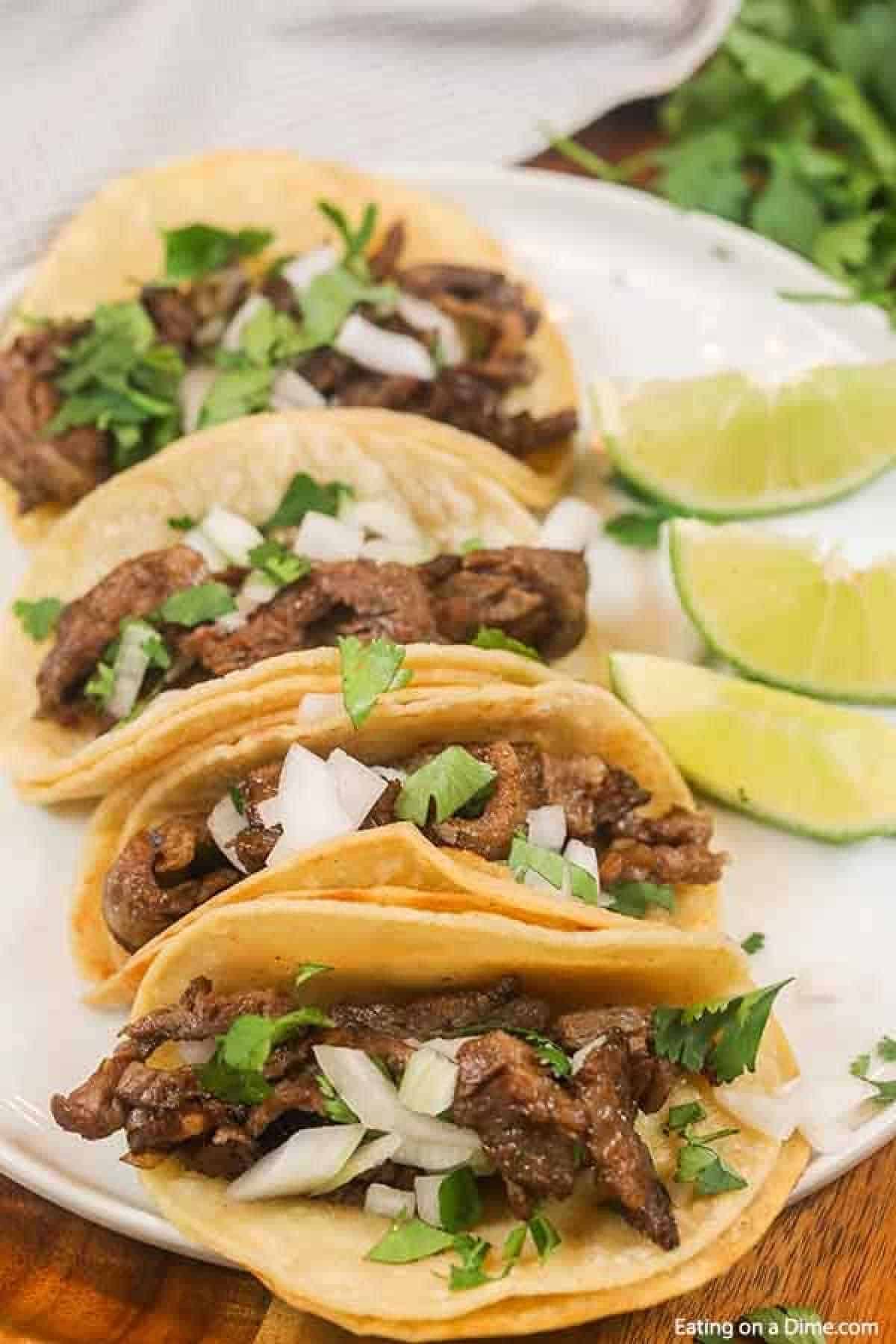 4 carne asada street tacos topped wit white onions and cilantro on a white plate with 3 lime wedges on the plate as well. 