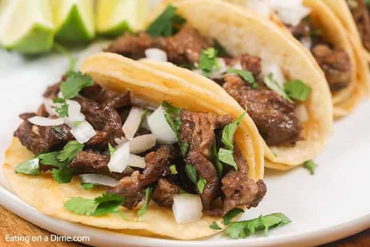 4 beef street tacos on a white plate topped with white onions and cilantro.  Each taco is on 2 corn tortillas. 