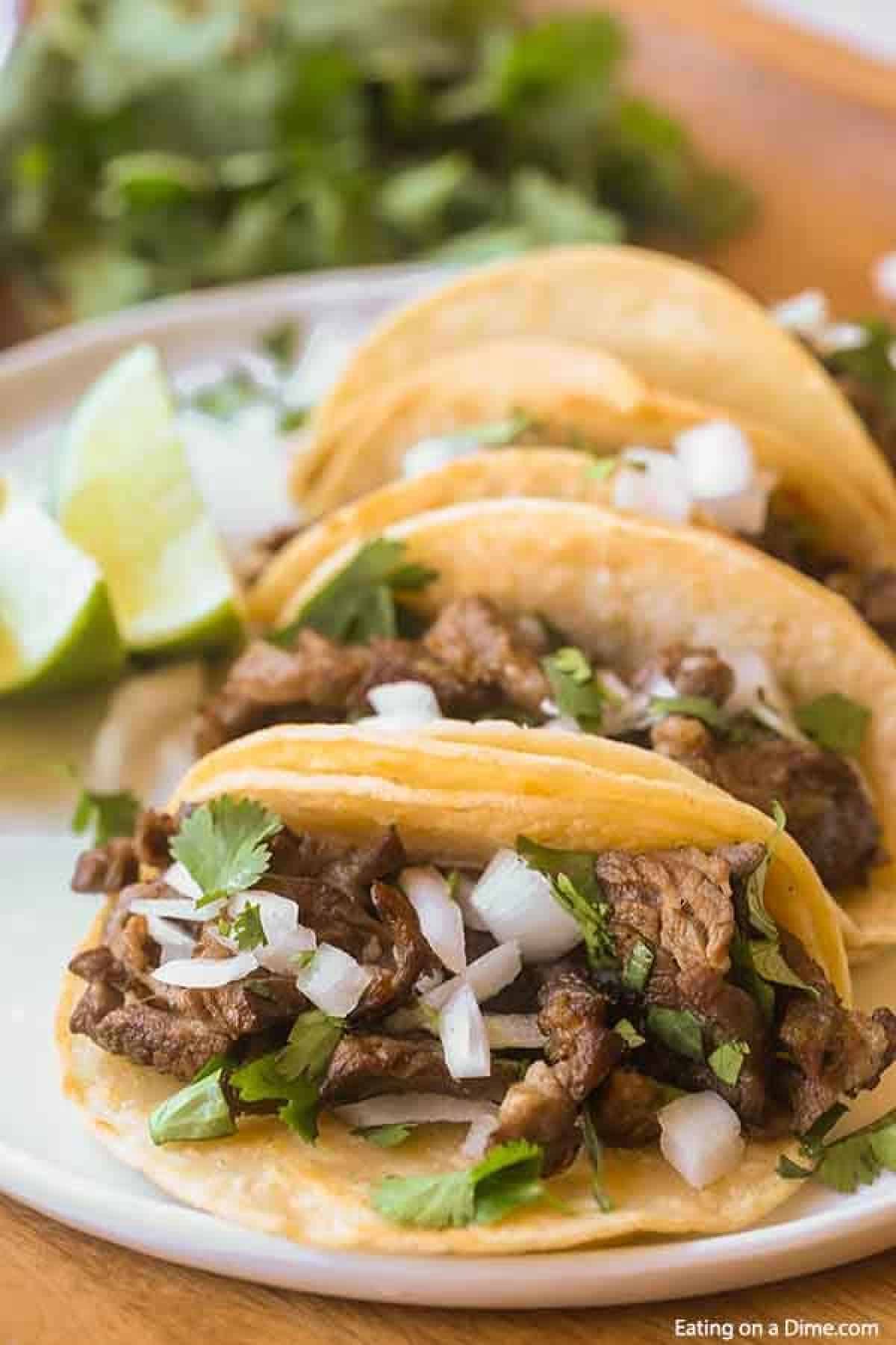 4 street tacos on a white plant with a bunch of cilantro behind them.  Each taco is topped with white onions and cilantro with lime wedges sitting next to the tacos. 