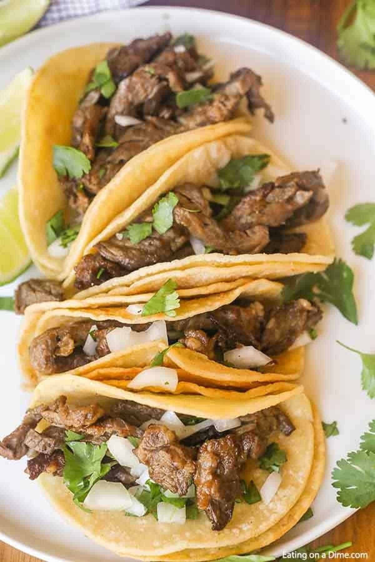 4 steak carne asada street tacos topped with white onions and cilantro. You can see lime wedges on the left side of the white plate that the tacos are on.