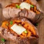 Two baked crock pot sweet potatoes split open, topped with pats of butter, chopped green onions, and a sprinkle of black pepper, displayed on a wooden surface.