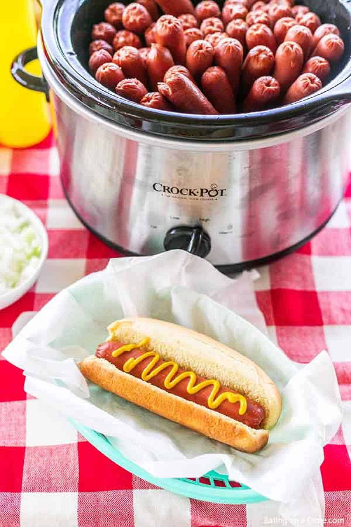 A hot dog with mustard on it in front of a small crock pot full of hot dogs 