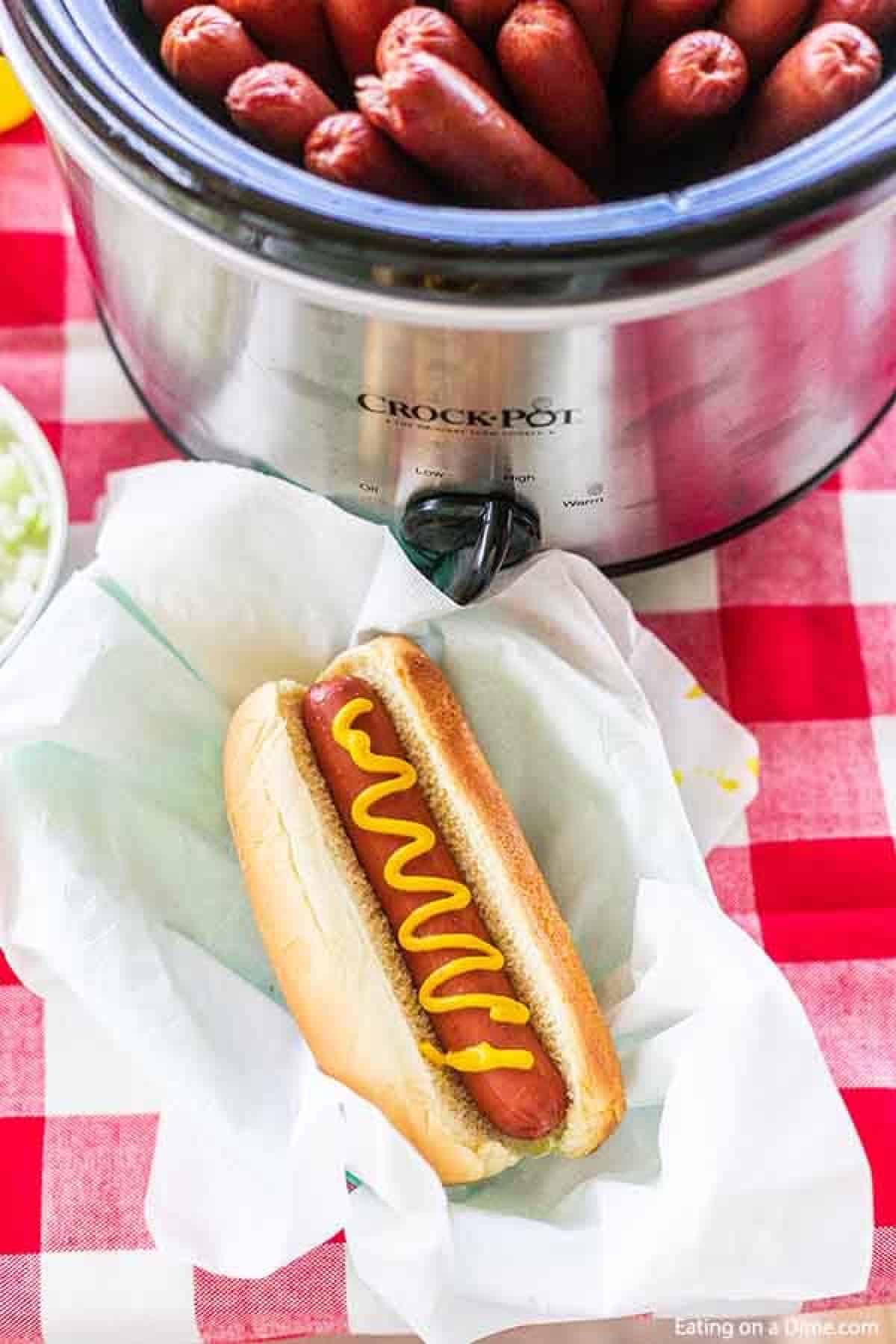 A hot dog with mustard on it in front of a small crock pot full of hot dogs 