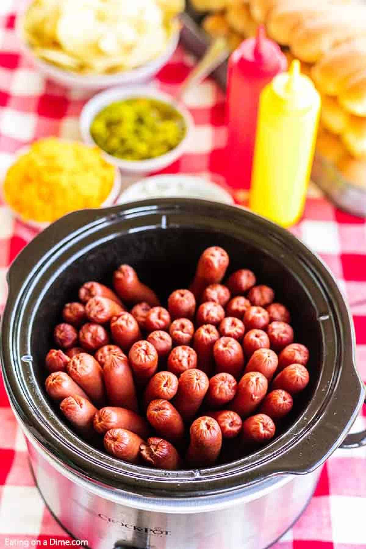 Crock Pot Hot Dogs are so easy and perfect for parties and to feed a crowd. The slow cooker does all of the work and the hot dogs stay warm. It is easy! Hot Dogs being cooked in a crock pot with other hot dog condiments behind it.