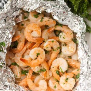 A pile of succulent grilled shrimp, seasoned with herbs, is served in a crumpled foil packet. Fresh parsley peeks through as a vibrant garnish around the dish.