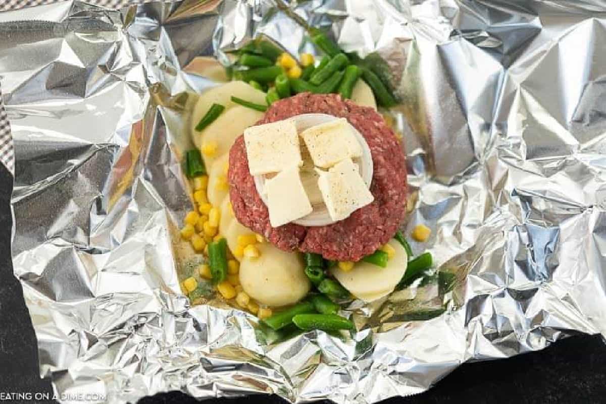 Piece of foil topped with green beans, corn, potatoes, hamburger patty, and butter slices