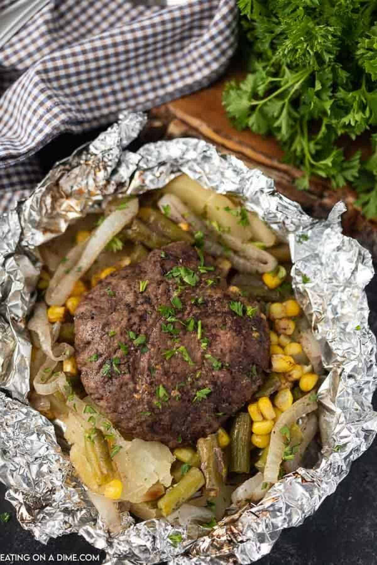 Hamburger patty in a piece of foil with slice onions, corn, and slice potatoes
