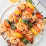 Grilled shrimp kabobs with zucchini, yellow squash, red bell pepper, and red onion are arranged on a white plate, with a lemon slice visible in the background.