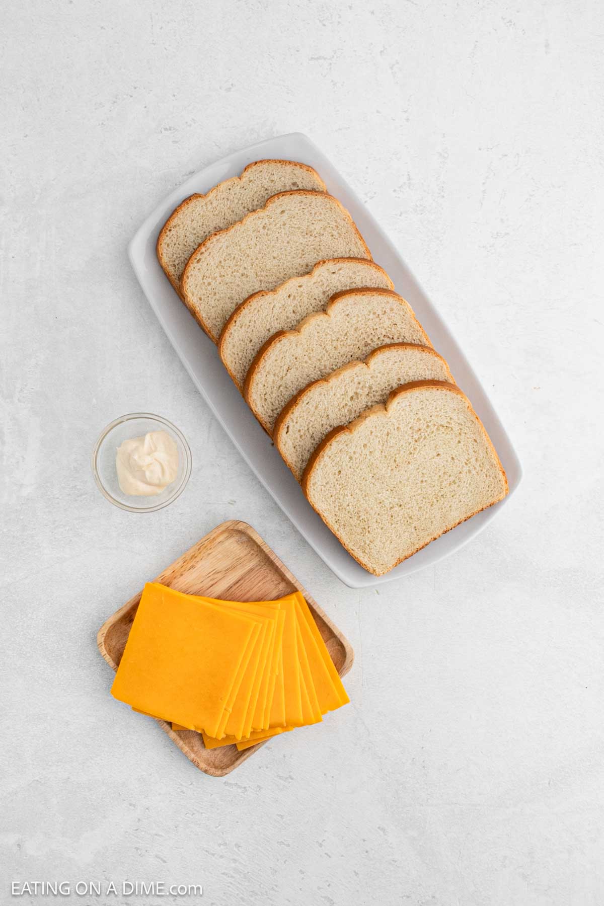 A white tray holds four slices of white bread, ready for transformation into a delicious grilled cheese sandwich. Nearby sits a glass dish of mayonnaise and a wooden plate with neatly arranged slices of orange cheddar cheese.