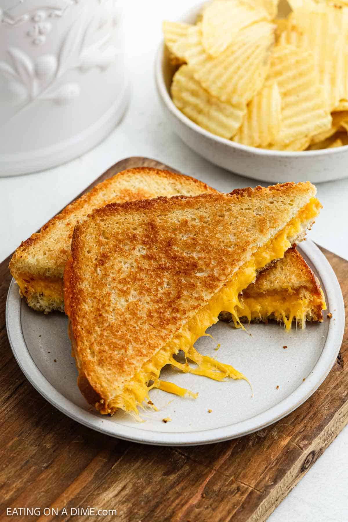 A plate showcases a grilled cheese sandwich cut diagonally, unveiling gooey melted cheese. This classic delight rests on a wooden board, accompanied by a bowl of crispy potato chips in the background.