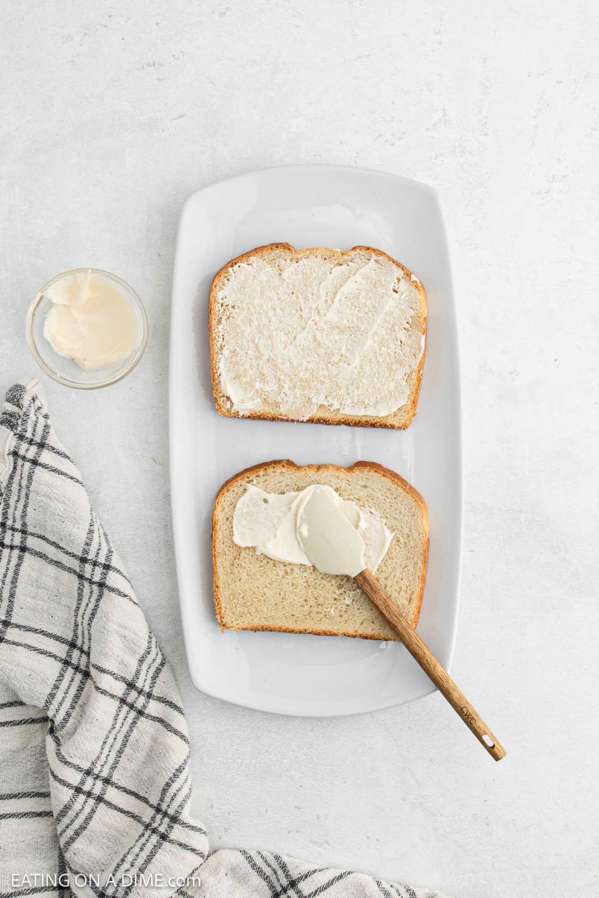 On a white plate, two slices of bread are topped with butter, ready to transform into a delicious grilled cheese sandwich. A knife rests on the bottom slice, gleaming with buttery promise. Nearby, a small glass jar of butter and a checkered cloth complete the cozy scene.