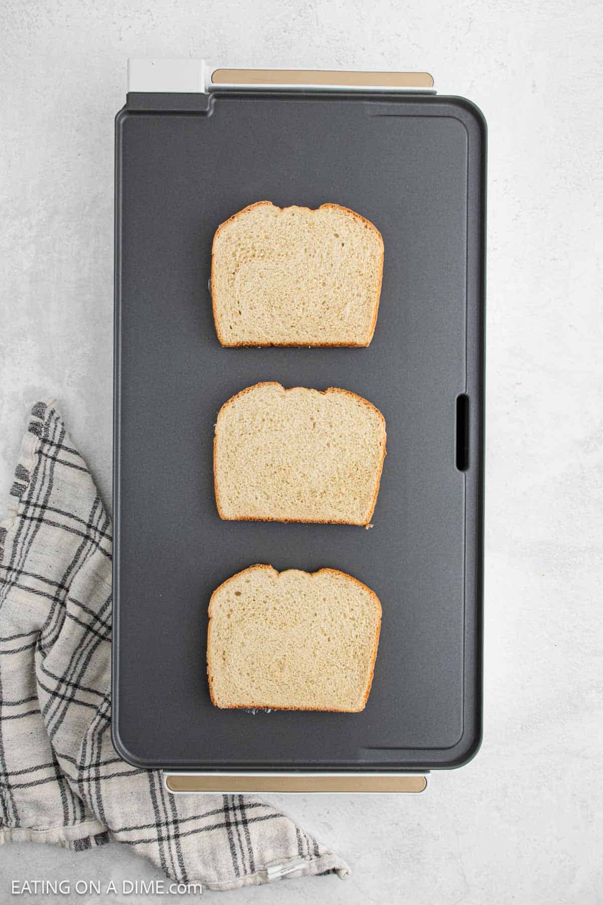 Three slices of bread are evenly spaced on a rectangular griddle, preparing for the perfect grilled cheese sandwich. A plaid kitchen towel is positioned beside the griddle on a light-colored surface.