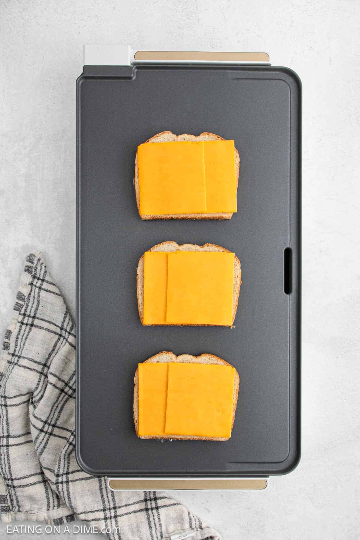Three slices of bread topped with cheddar cheese sizzle on the griddle, crafting a perfect grilled cheese sandwich. A black and white checkered cloth peeks out from beside the griddle.