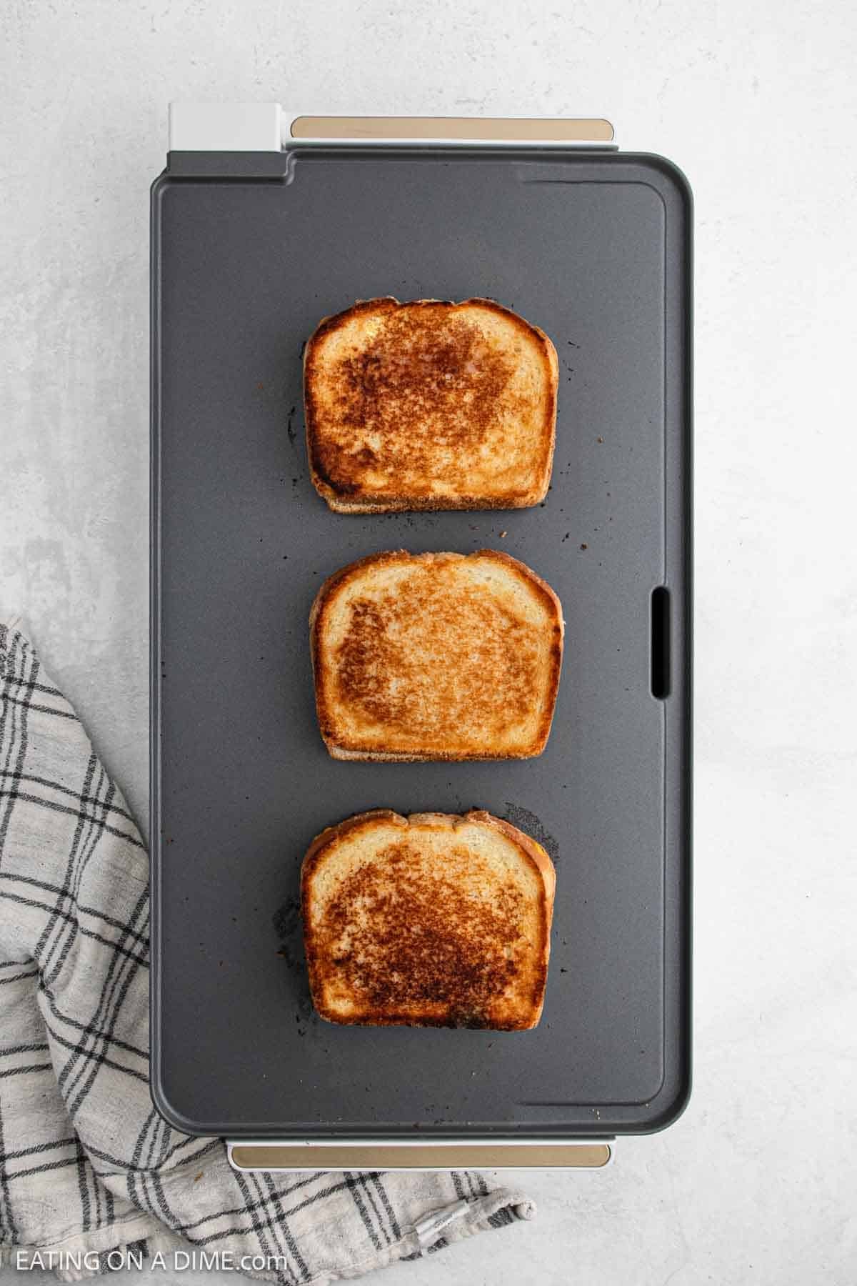 Three golden-brown grilled cheese sandwiches are aligned vertically on a rectangular griddle. Neatly arranged, they rest against a backdrop where a white and black checkered cloth peeks into view on the left side.
