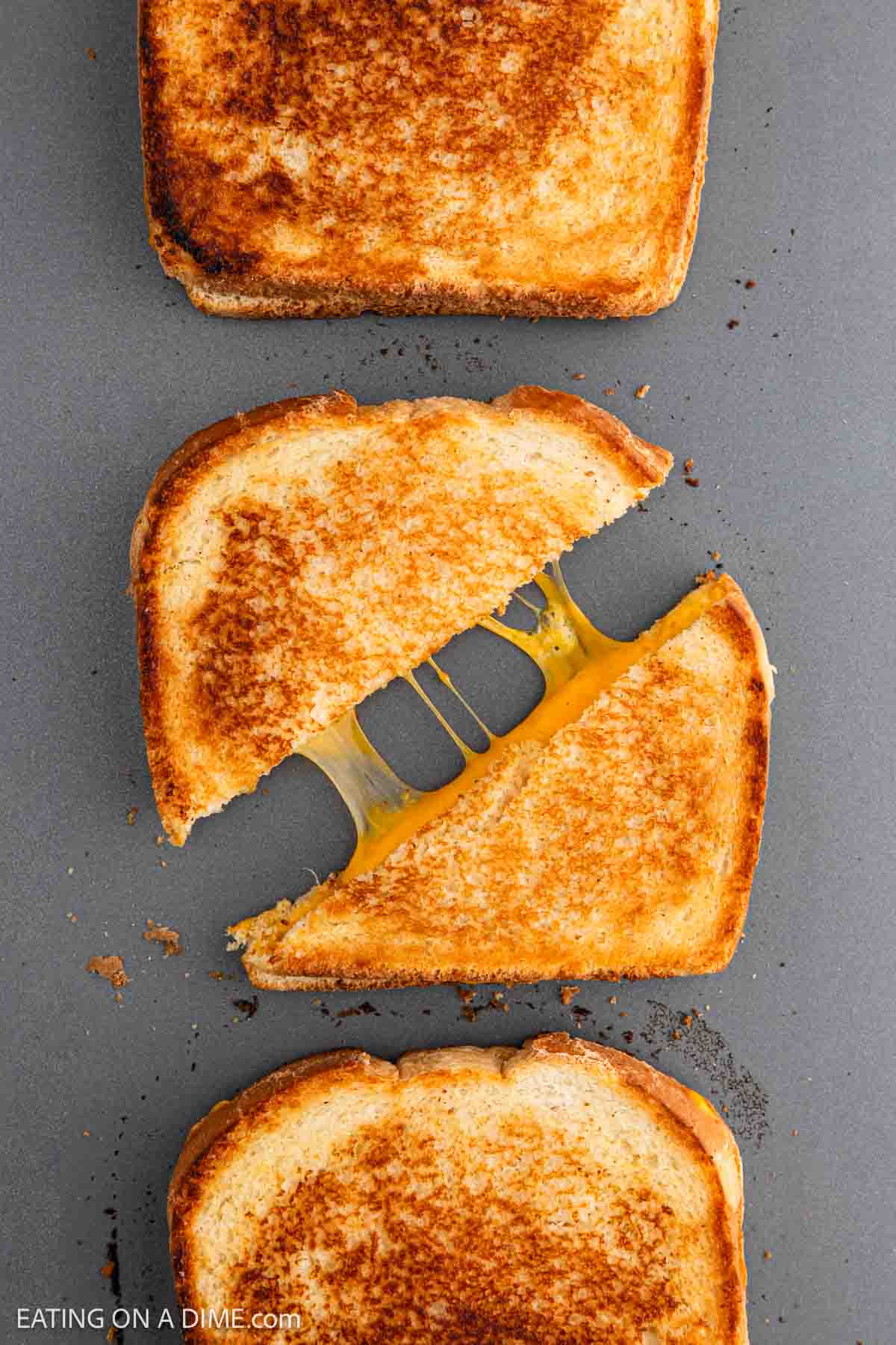 Three grilled cheese sandwiches rest on a gray surface. A sandwich cut diagonally reveals gooey melted cheese perfectly stretched between the halves, inviting you to savor every bite.