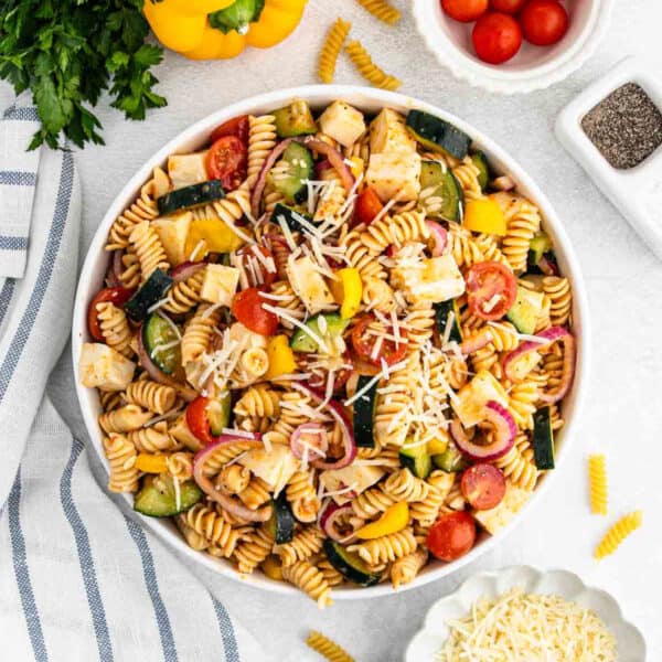 A bowl of Salad Supreme Pasta Salad with rotini, cherry tomatoes, yellow bell peppers, cucumbers, red onions, cheese, and grated parmesan, surrounded by fresh ingredients and a striped towel.