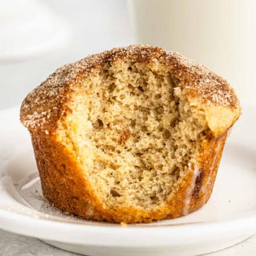 A Snickerdoodle Muffin with a bite taken out sits on a white plate. The muffin has a soft, airy texture and a lightly golden brown top sprinkled with cinnamon sugar for that classic snickerdoodle flavor.