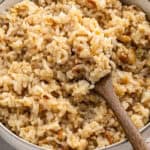 A close-up of a bowl of Stick of Butter Rice with caramelized onions, being stirred with a wooden spoon. The rice looks fluffy and slightly browned.